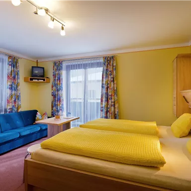 A bright room with a double bed and two yellow pillows. There is a blue sofa, a table, and large windows with colorful curtains.
