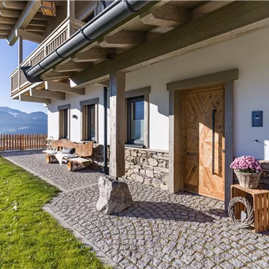 An inviting entrance area of a house with a stone path and a wooden veranda. In the background, gentle hills and a blue sky can be seen.
