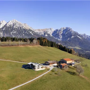 A picturesque alpine landscape with snow-covered mountains in the background. In the foreground lies a farmhouse on green meadows.