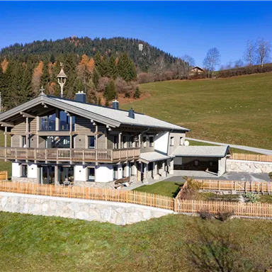 A modern wooden house in an idyllic landscape with green meadows and gentle hills. In the background, there are trees and a clear blue sky.
