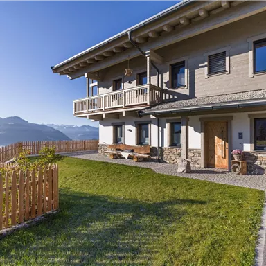 A modern wooden house with a balcony and a well-kept garden. In the background, mountains and a clear sky can be seen.