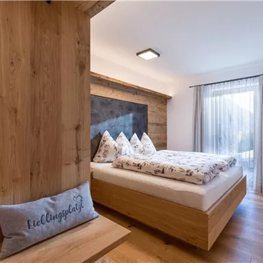 A modern bedroom with a cozy bed and wooden furniture. Large windows provide plenty of daylight and a view outside.