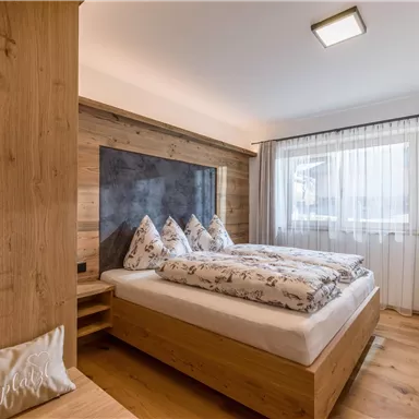 A modern bedroom with a cozy double bed and wooden details. Large windows provide plenty of daylight.