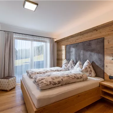 A modern bedroom with a double bed and wooden furniture. Large windows provide plenty of daylight and a view of the outdoors.