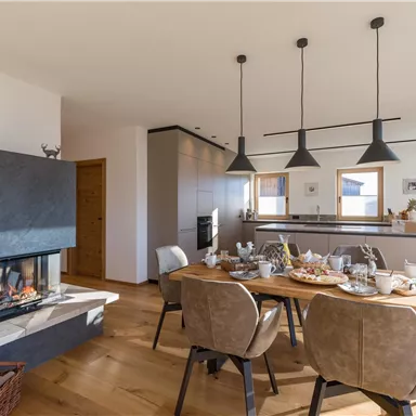 A modern dining room with a cozy fireplace and stylish furniture. The table is set, and there is plenty of daylight coming through the large windows.
