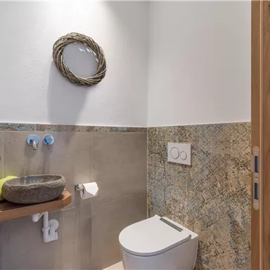 A modern bathroom with a natural stone sink and a toilet. A decorative wreath hangs on the wall, and the tiles are in earth tones.