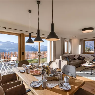 A modern living room with large windows and a view of the mountains. The room is cozily furnished with a dining area and an inviting seating group.