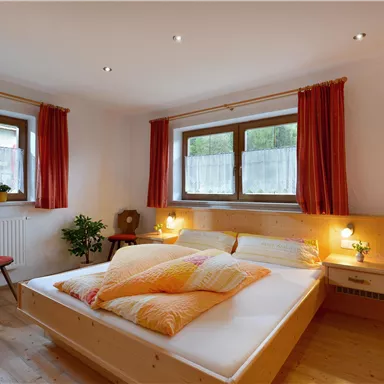 Ein gemütliches Schlafzimmer mit einem großen Bett und bunten Kissen. Die Fenster lassen viel Tageslicht herein und die orangefarbene Gardinen sorgen für eine warme Atmosphäre.