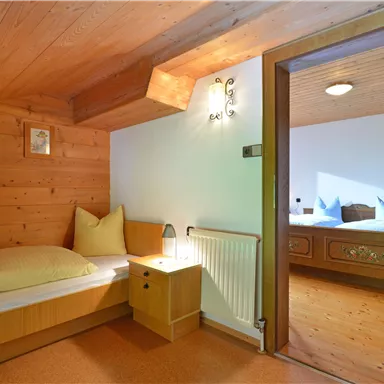 Ein gemütliches Schlafzimmer mit Holzverkleidung und einem Einzelbett. Im Hintergrund ist ein weiterer Raum mit zwei Betten sichtbar.