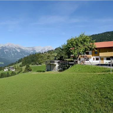 Ein malerisches Haus auf einer grünen Wiese, umgeben von Bergen. Der Himmel ist klar und die Landschaft ist idyllisch.