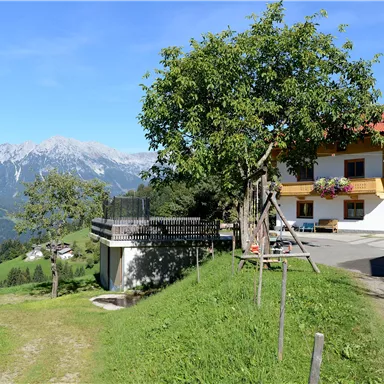 Ein ruhiges ländliches Anwesen mit einem Wohnhaus und einem großen Baum. Im Hintergrund sind majestätische Berge und eine klare blaue Himmel zu sehen.