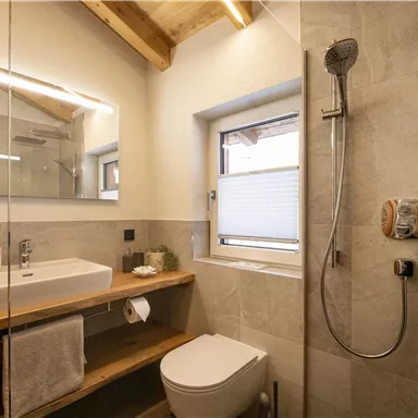 Ein modernes Badezimmer mit einer Dusche, einem Waschtisch aus Holz und einer großen Spiegelfläche. Der Raum hat helle Fliesen und ein Fenster für natürliches Licht.