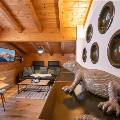 Eine gemütliche Wohnstube mit Holzdecken, modernen Möbeln und einer TV-Anlage. Auf einem Tisch liegt eine dekorative Echse und an der Wand hängen runde Wanddekorationen.