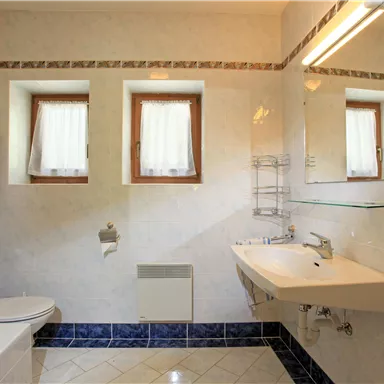 Ein helles Badezimmer mit Dusche, Waschbecken und WC. Fenster mit Vorhängen lassen Licht herein und sorgen für eine freundliche Atmosphäre.