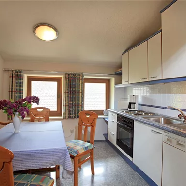 Eine helle Küche mit einem Esstisch und Stühlen. Auf dem Tisch steht eine Vase mit Blumen und die Fenster sind mit Vorhängen dekoriert.