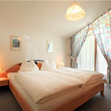 Ein gemütliches Schlafzimmer mit einem großen Doppelbett und blühenden Vorhängen. Es gibt sanftes Licht von den Tischlampen und einen Blick auf den Außenbereich durch die Fenster.