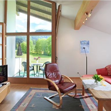 Ein modernes Wohnzimmer mit großen Fenstern und Blick auf die grüne Landschaft. Die Möbel sind gemütlich, einschließlich eines roten Sofas und eines Holzkaffee­table.