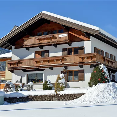 Ein wunderschönes Chalet im Schnee mit einem Holzbalon und grünen Sträuchern. Der klare blaue Himmel ergänzt die winterliche Szenerie.