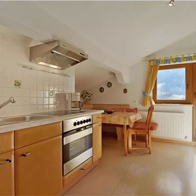 Eine moderne Küche mit Holzschränken und ausgestattet mit einem Herd, Spülbecken und einer Essecke. Große Fenster sorgen für viel Tageslicht.