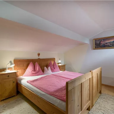 Ein gemütliches Schlafzimmer mit einem Holzbett und rosa Kissen. Neben dem Bett stehen Nachttische und ein Stuhl in einem hellen Raum.