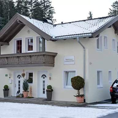 A modern two-story house with a balcony and a snow-covered roof. In front of the house, there are two planters and a black car.