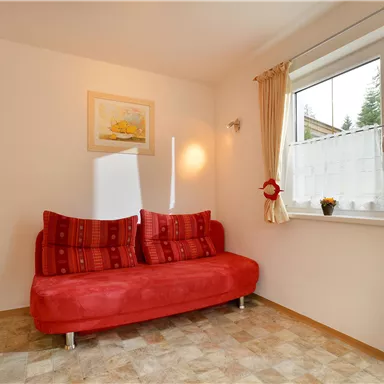 A cozy room with a red sofa and a window that lets in a lot of light. A picture hangs on the wall, and a small flower is on the windowsill.