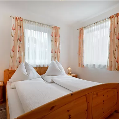 A cozy bedroom with a wooden bed and white bedspreads. Large windows with curtains let in plenty of light.