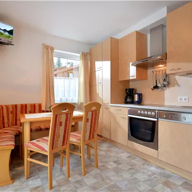 A modern kitchen with a dining table and wooden chairs. The walls are light, and there are large windows that let in plenty of light.