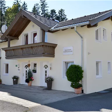 A modern, bright house with a balcony and surrounded by green trees. The facade is in a light yellow tone.