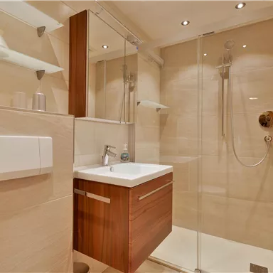 Ein modernes Badezimmer mit einer Glasdusche und einem schlichten Waschtisch aus Holz. Die Wände sind in hellen, neutralen Farben gehalten.