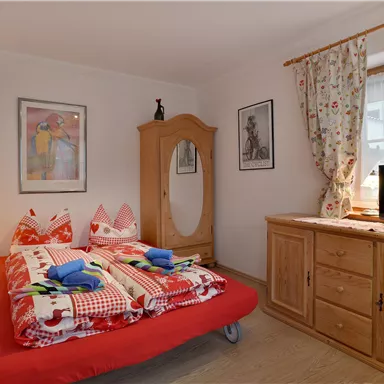 Ein gemütliches Schlafzimmer mit einem roten Bett, bunten Kissen und einem Holzschrank. Ein Fernseher steht auf einer Kommode, und die Fenster sind mit Vorhängen geschmückt.