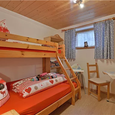 Ein gemütliches Schlafzimmer mit einem Etagenbett und bunten Kissen. Ein kleiner Tisch und ein Stuhl ergänzen den Raum, der von natürlichem Licht durch ein Fenster erhellt wird.