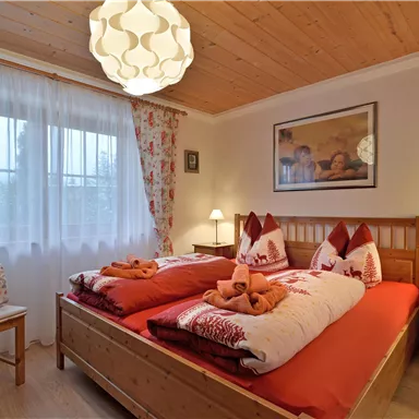 Ein gemütliches Schlafzimmer mit zwei Betten, roter Bettwäsche und dekorativen Kissen. Das Zimmer ist hell und freundlich mit einem großen Fenster und einer modernen Lampe.