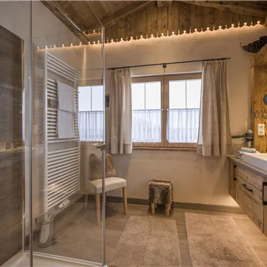 Ein modernes Badezimmer mit einer gläsernen Dusche und einem Holzmobiliar. Große Fenster sorgen für viel Licht und eine angenehme Atmosphäre.