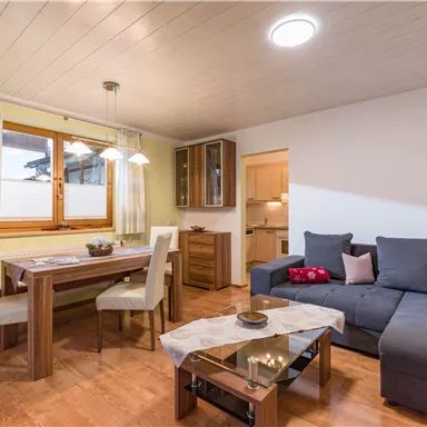 Ein modernes Wohnzimmer mit einem bequemen grauen Sofa und einem Esstisch aus Holz. Große Fenster lassen viel Licht herein und schaffen eine gemütliche Atmosphäre.