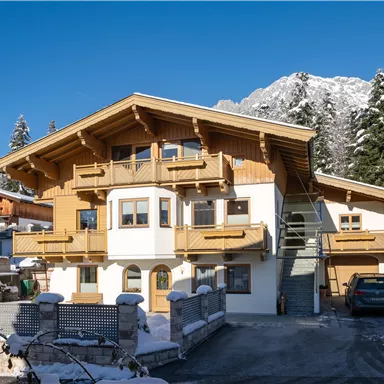 Ein schönes, traditionelles Alpine-Haus mit Holzelementen. Im Hintergrund sind schneebedeckte Berge und grüne Nadelbäume zu sehen.