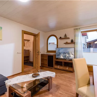 Ein gemütliches Wohnzimmer mit modernen Möbeln. Es gibt ein Sofa, einen Fernseher und große Fenster, die viel Licht hereinlassen.