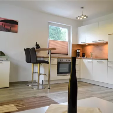 A modern kitchen with white cabinets and wooden details. In the background, there is a TV and a picture of a landscape.