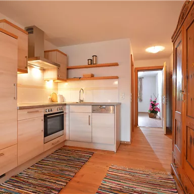 Eine moderne Küche mit hellen Holzschränken und einem freundlichen Design. Im Hintergrund ist ein Flur mit Blumen zu sehen.