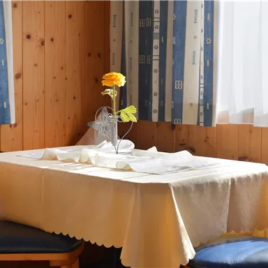 Ein gemütlicher Tisch mit einer weißen Tischdecke in einem Holzraum. In der Mitte steht eine blühende Blume in einer Vase.