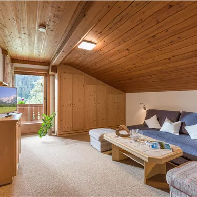 Ein gemütlicher Wohnraum mit holzverkleidetem Dach und Blick auf die Berge. Die Einrichtung besteht aus einem grauen Sofa, einem Holztisch und einem Fernseher.