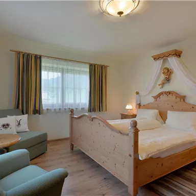 A cozy bedroom with a wooden bed and decorative details. A small seating area and daylight through the window create an inviting atmosphere.