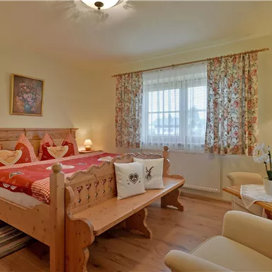 A cozy bedroom with a double bed and light wooden furniture. The room is cheerfully decorated and has large windows with floral curtains.