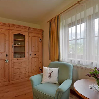 A cozy room with a wooden display cabinet and a bright armchair. The windows are adorned with curtains, and a small table with fresh flowers stands next to it.