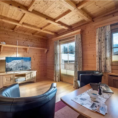 Ein gemütlicher Wohnraum mit Holzdecken und warmen Farben. Im Vordergrund stehen zwei schwarze Sessel und ein Tisch mit Zeitschriften, im Hintergrund ist ein Fernseher sichtbar.