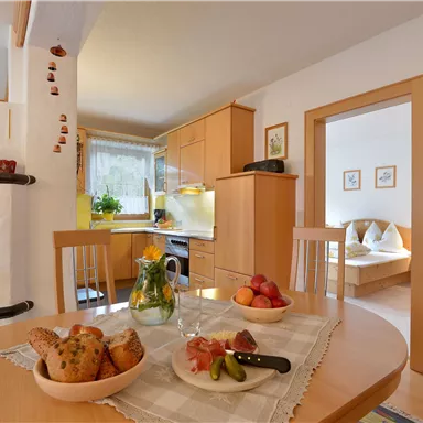 Eine gemütliche Küche mit einem Holztisch und frischem Obst auf einem Tisch. Im Hintergrund sind Küchenmöbel und ein angrenzendes Zimmer sichtbar.