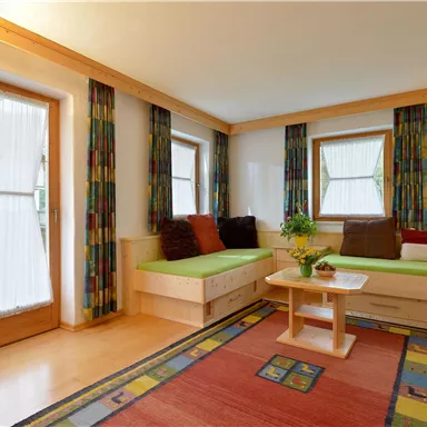 Ein helles Wohnzimmer mit gemütlichen Sitzgelegenheiten und großen Fenstern. Der Raum ist mit bunten Vorhängen und einem Teppich dekoriert.