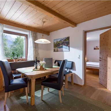 Ein gemütlicher Essbereich mit einem Holztisch und schwarzen Stühlen. Im Hintergrund sind Fenster mit Ausblick und ein weiterer Raum sichtbar.