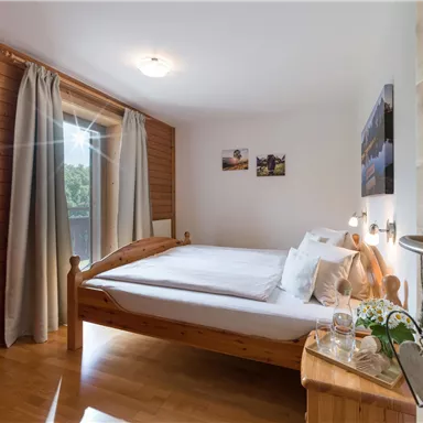 Ein helles Schlafzimmer mit einem großen Bett und Holzverkleidungen. Vorhänge lassen Licht herein, und es gibt einige dekorative Bilder an der Wand.