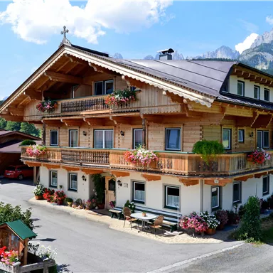 Ein traditionelles alpine Gasthaus mit einem Holzbau und bunten Blumen auf den Balkonen. Im Hintergrund sind majestätische Berge und ein blauer Himmel sichtbar.